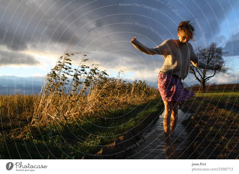 Woman standing in puddle in field Young woman Skirt Feminine Day Field Meadow acre Puddle Clouds Storm clouds windy Nature Landscape Barefoot somber Sunlight