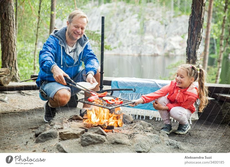 Man and his little daughter having barbecue in forest on rocky shore of lake, making a fire, grilling bread, vegetables and marshmallow. Family exploring Finland. Scandinavian summer landscape.
