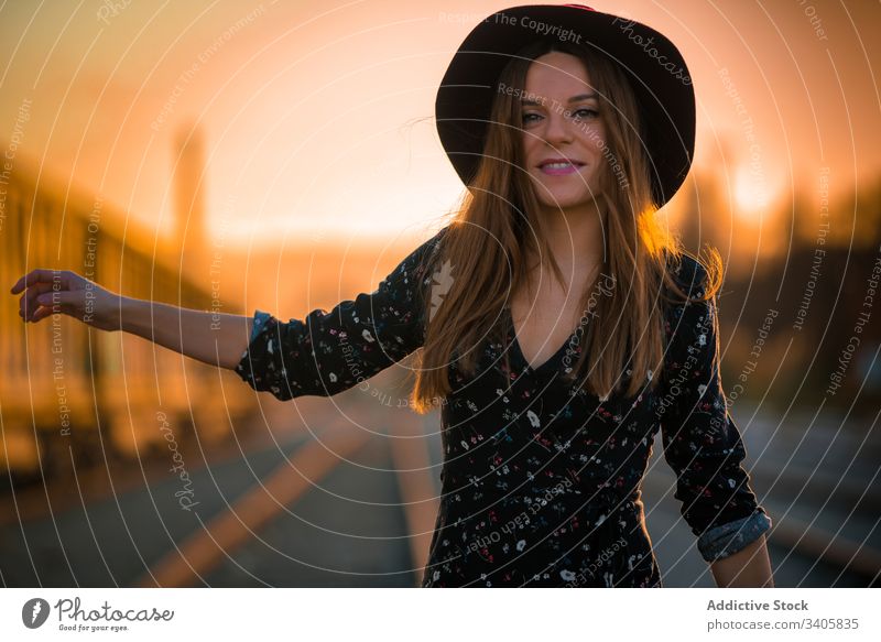 Cheerful woman hitchhiking on railroad at sunset hitchhike countryside smile gesture travel style female hat lifestyle trip route way highway twilight dusk