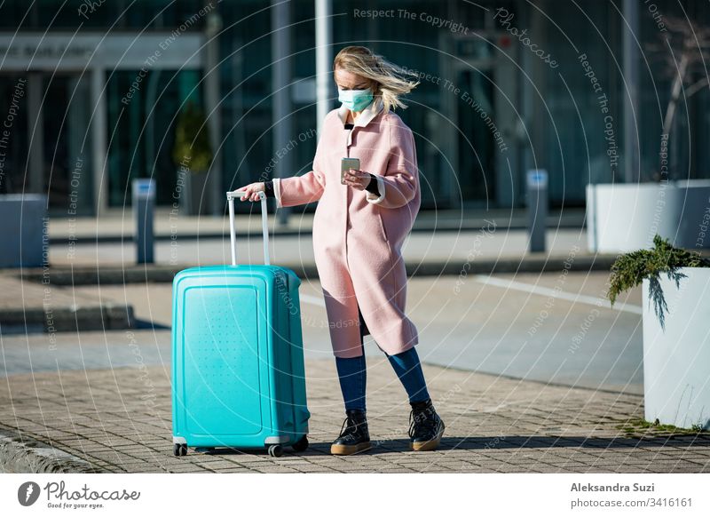 Woman in protective mask standing at airport entrance with smartphone and big luggage case, browsing, texting, using mobile app. Safe traveling. arrival