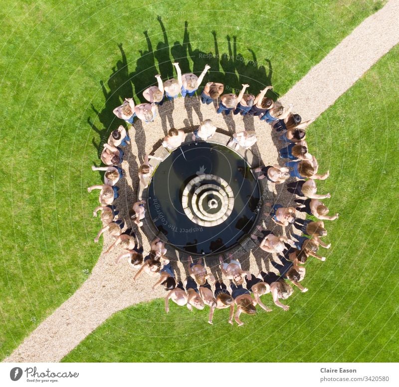 Aerial view of about 50 women in jeans, around an ornamental fountain, with a diagonal path and green surround. Breast awareness sisters breast cancer
