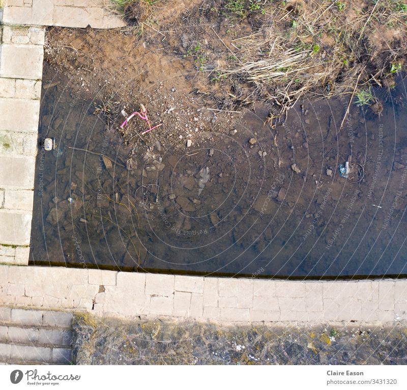 Aerial image of a childs' abandoned pink scooter, lying at edge of a shallow stream,  partly framed by a stone edging. lonely Loneliness Grief Sadness Distress