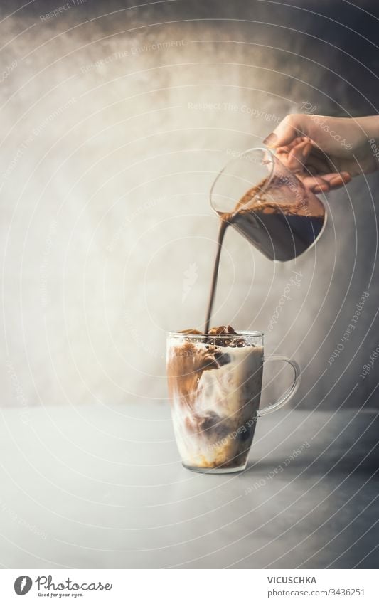Barista hand pouring coffee cream in glass mug of iced coffee latte on rustic table at concrete wall background. Iced coffee making. Summer refreshing beverage. Cold drink. Ice coffee preparation.