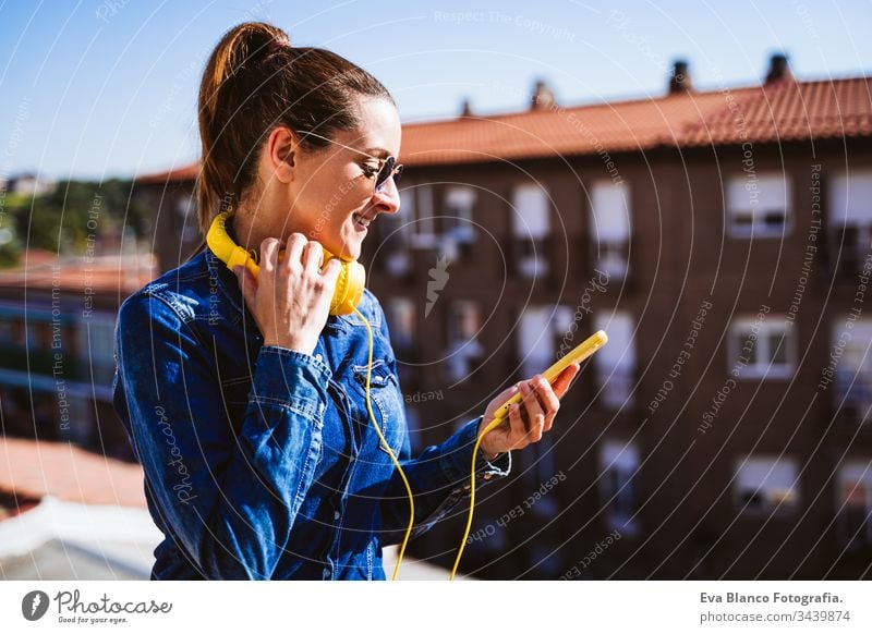 young woman listening to music on mobile phone and yellow headset. Fun and lifestyle technology fun terrace outdoors sunny sunglasses happy caucasian active
