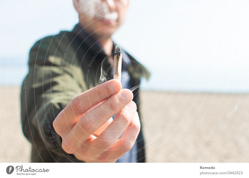 Young man holding a lit marijuana joint while smoking on the beach. Blur background and copy space right. person cannabis smoke outdoor weed casual lifestyle
