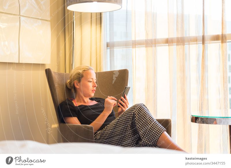 Businesswoman on business travel, tired from business meeting, sitting alone in hotel room afterwork, reading and responding to emails and messages on her smart phone