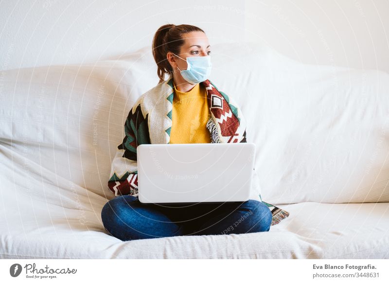 young woman working on laptop at home, sitting on the couch, wearing protective mask. Stay home concept during coronavirus covid-2019 pandemic stay home