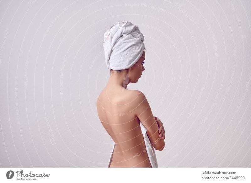 Skinny young woman after the shower from her back with white towel over her head, and with bare back. Half-length portrait against plain light grey background