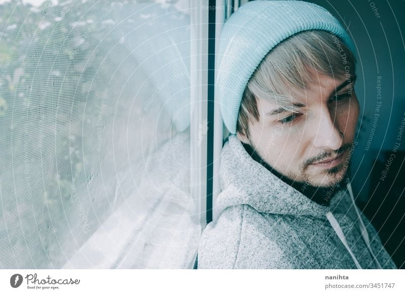 Young man alone in his home near a window male portrait isolated locked quarantine lonely self attractive handsome guy casual wear hairstyle blue eyes lenses