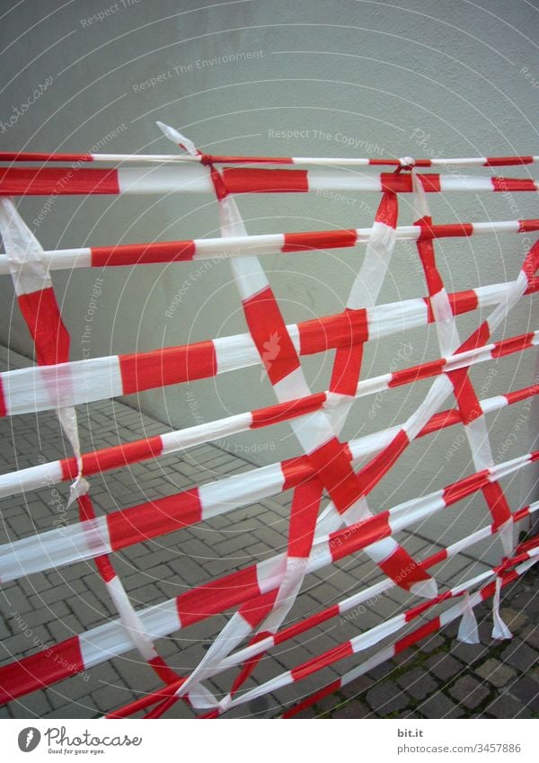 Barrier tape, hangs criss-cross, across a street, in front of a grey house wall. Protection against infection with Corona during the Corona Pandemic. Tape for protective measures, hygiene regulation, distance rule, minimum distance, corona determination, infection with virus