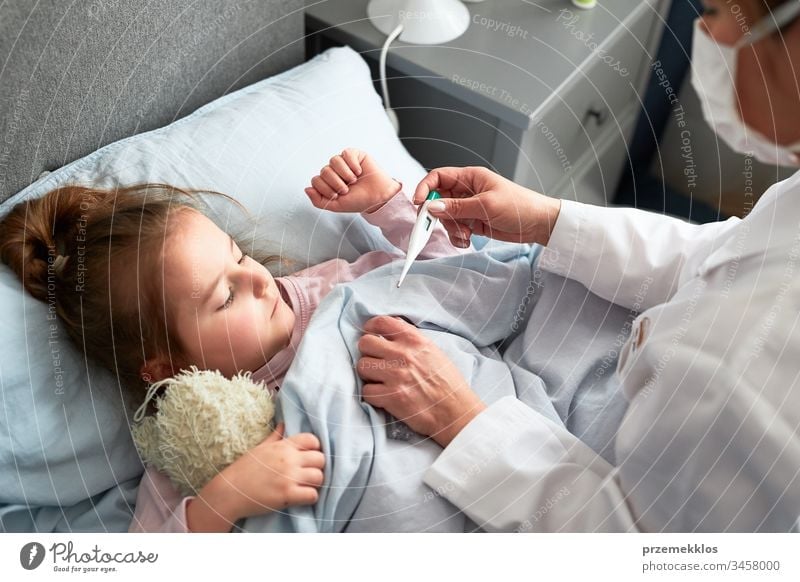 Doctor visiting little patient at home. Measuring the temperature of sick girl lying in bed. Woman wearing uniform and face mask. Medical treatment child virus