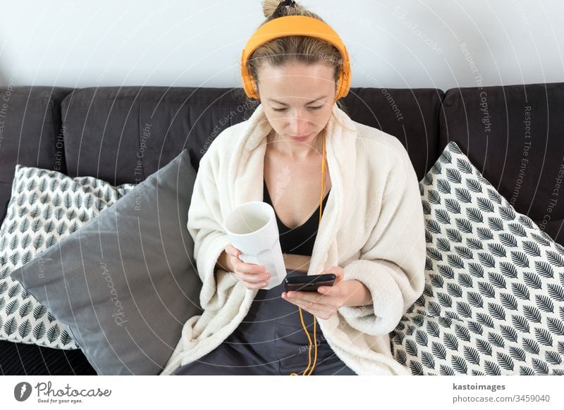 Stay at home. Social distancing. Woman at home relaxing on sofa couch drinking tea from white cup, listening to relaxing music, stay connected to friens and family via social networks on mobile.