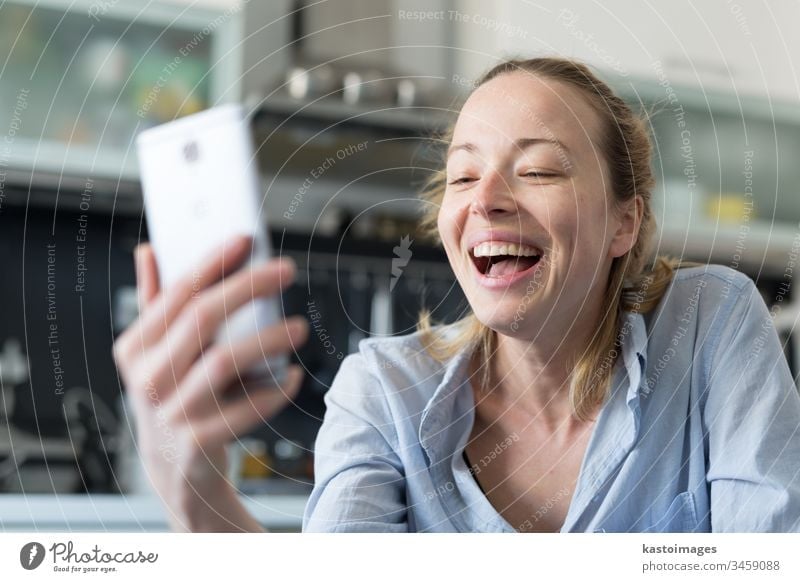 Young smiling cheerful pleased woman indoors at home kitchen using social media apps on mobile phone for chatting and stying connected with her loved ones. Stay at home, social distancing lifestyle.