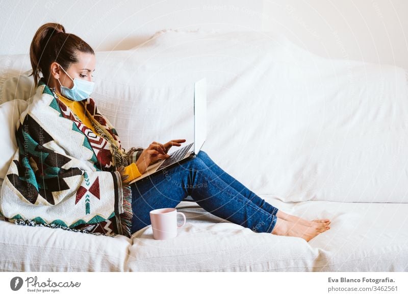 young woman working on laptop at home, sitting on the couch, wearing protective mask. Stay home concept during coronavirus covid-2019 pandemic stay home