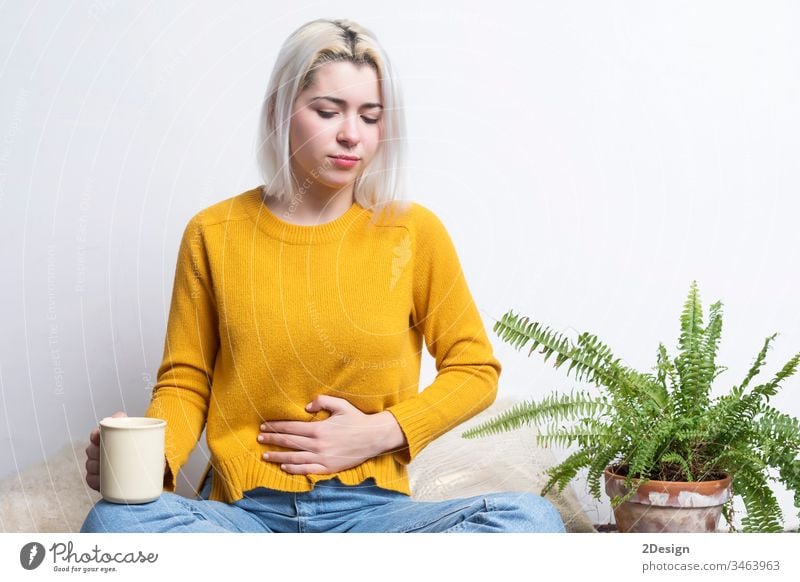Young blonde female sitting at home holding a cup of tea while putting her hand in her stomach health care unhealthy menses medicals medicine sick hot drink ill