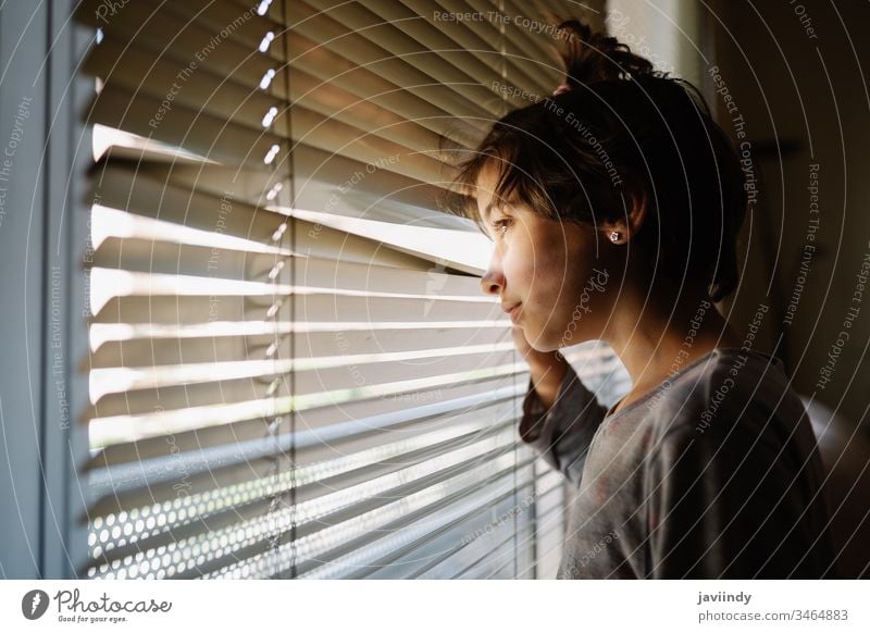 Nine-years-old girl looking out the window through the blinds stay at home young female quarantine person face day room caucasian beautiful portrait house