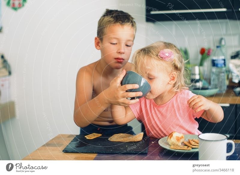 Elder brother and little sister having breakfast in the kitchen. Careful boy helping girl drink from the cup children morning care tea meal siblings kid eat
