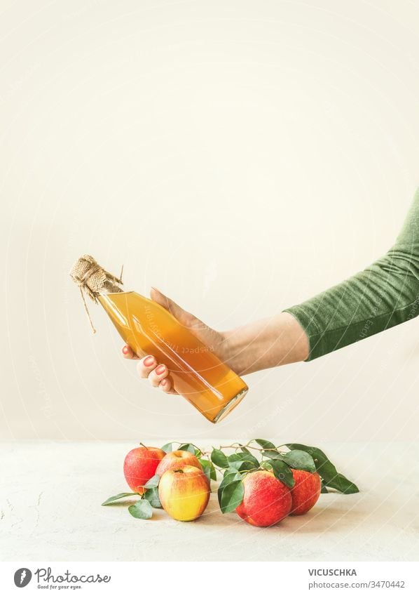 Female women hand holding bottle with homemade apple cider vinegar above table with apples and green leaves at white wall background. Apple season. Healthy fermented food concept