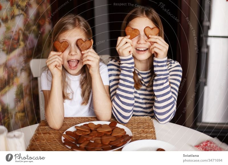 Charming, funny and laughing girls hold heart-shaped cookies, close their eyes and fool around. Valentine's Day and Women's Day cute kitchen female baby bakery