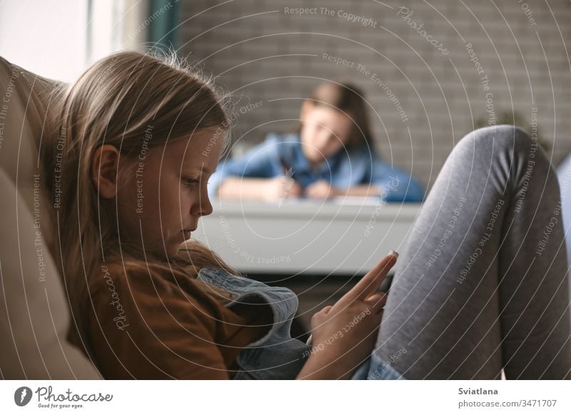 A cute girl plays on her phone while her sister does her homework. Communication, social distance during quarantine. Distance learning, online education