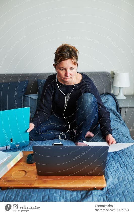 Woman working doing her job remotely during video chat call stream online course webinar on laptop from home. Woman sitting on bed in front of computer looking at screen using headphones and smartphone