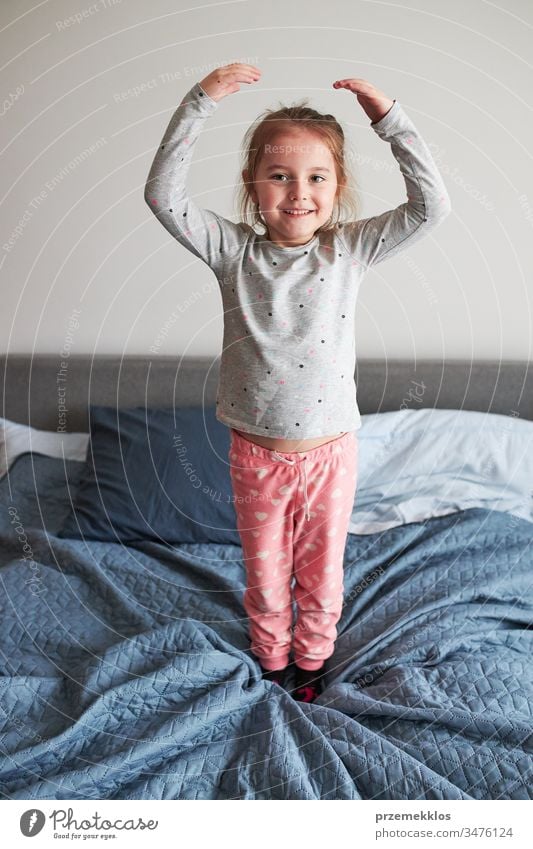 Little happy playful kid girl having fun making silly funny faces jumping on bed in bedroom in the morning at home playing childhood joy cute laughing smiling