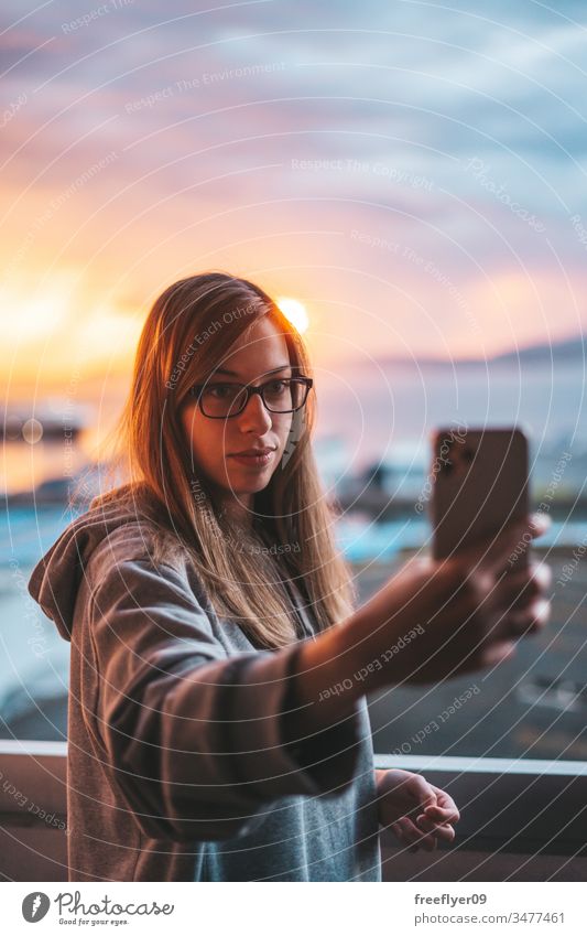 Young woman taking a selfie with the ocean on the background smartphone photograph photographing sunset balcony quarantine grey sweatshirt blonde glasses young