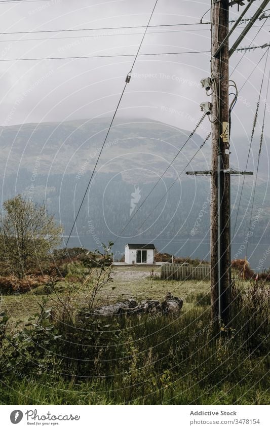 Lonely house at lakeside in mountainous terrain with electric cables countryside pole overcast fog rural landscape scotland gloomy cottage shore water highland