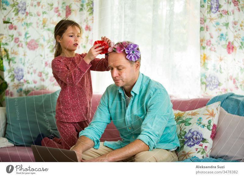 Child playing and disturbing father working remotely from home. Little girl combing daddy's hair and making hairstyle. Man sitting on couch with laptop. Family spending time together indoors.