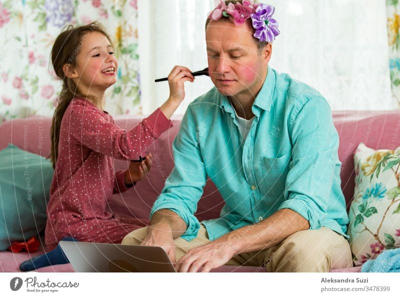 Child playing and disturbing father working remotely from home. Little girl applying makeup with brush. Man sitting on couch with laptop. Family spending time together indoors.