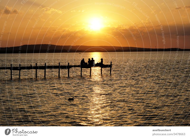 sunset over lake Steinhuder Meer in Germany silhouette sea pier jetty group friends unrecognizable people sitting dock watching sundown evening outdoor