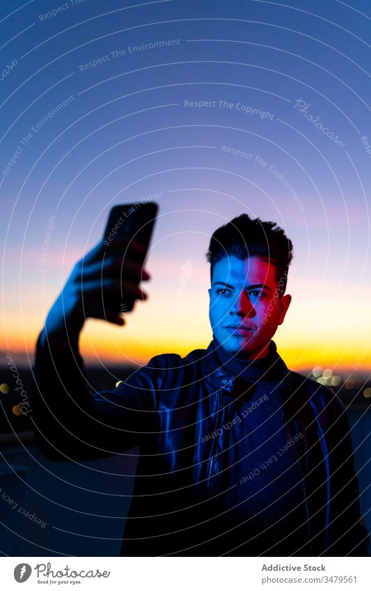 Young man taking selfie in evening time smartphone neon mobile phone sunset dark blue light sky style serious color gadget device modern trendy confident young