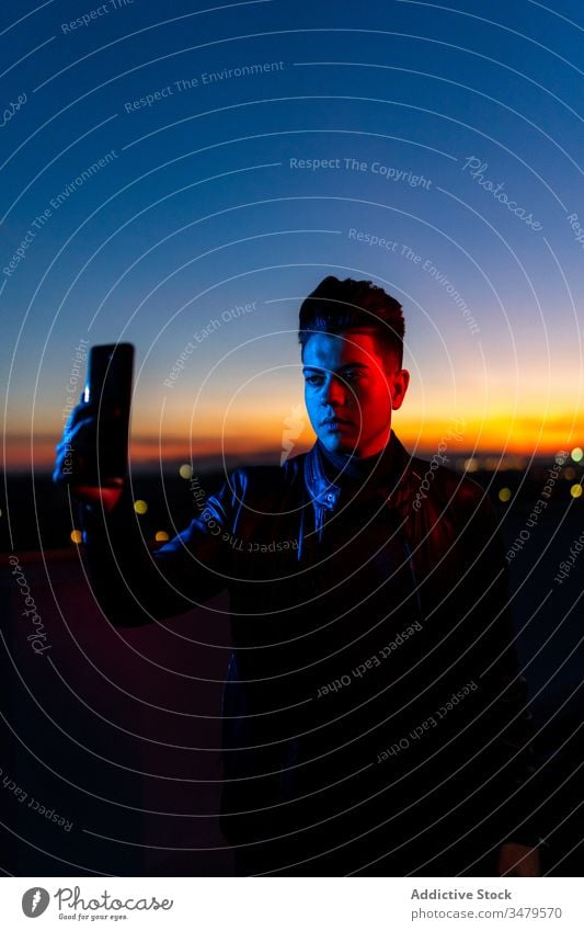 Young man taking selfie in evening time smartphone neon mobile phone sunset dark blue light sky style serious color gadget device modern trendy confident young