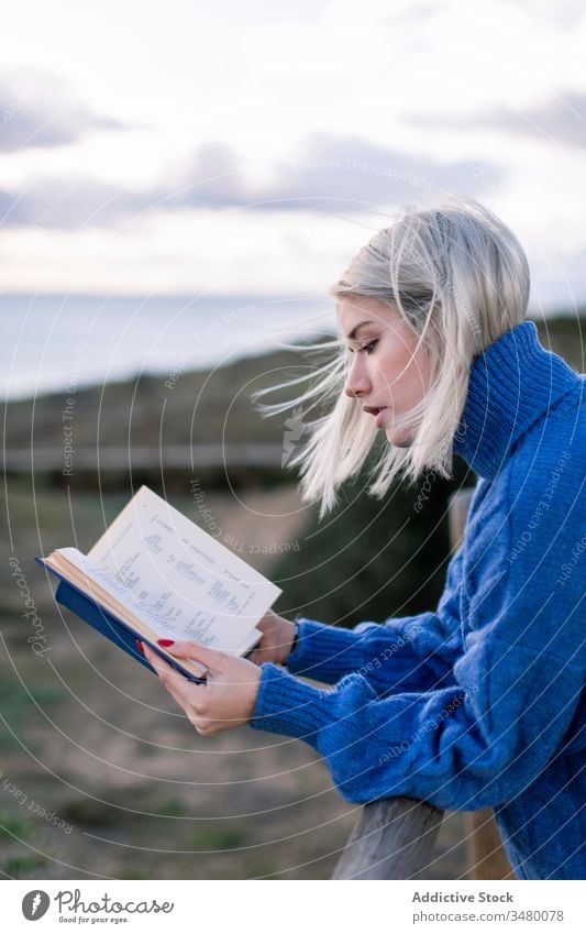 Young woman reading book near sea fence beach rest young blue enjoy poetry sweater nature relax female wooden style alone modern trendy blond calm seaside