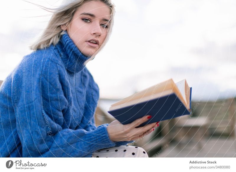 Young woman reading book on seashore terrace bench beach rest young blue sweater nature relax female wooden style vacation enjoy holiday modern trendy blond