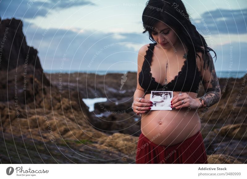 Loving stylish pregnant woman with sonogram picture on shore nature love anticipate parent expect freedom harmony sky sensual style coast travel prenatal