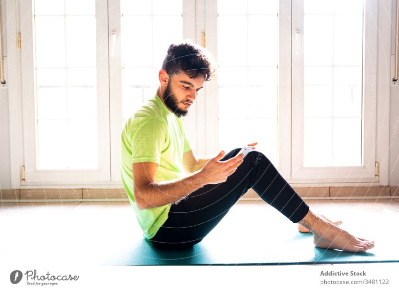 Pensive relaxed athlete using smartphone while resting after training on sports mat in light gym sportsman messaging use browsing watch read search surfing home
