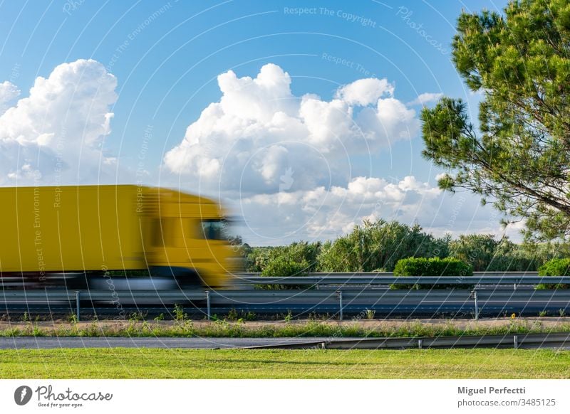 Yellow truck in motion driving on the highway next to a tree and a blue sky with clouds. trailer cargo road field landscape transport lorry fog foggy