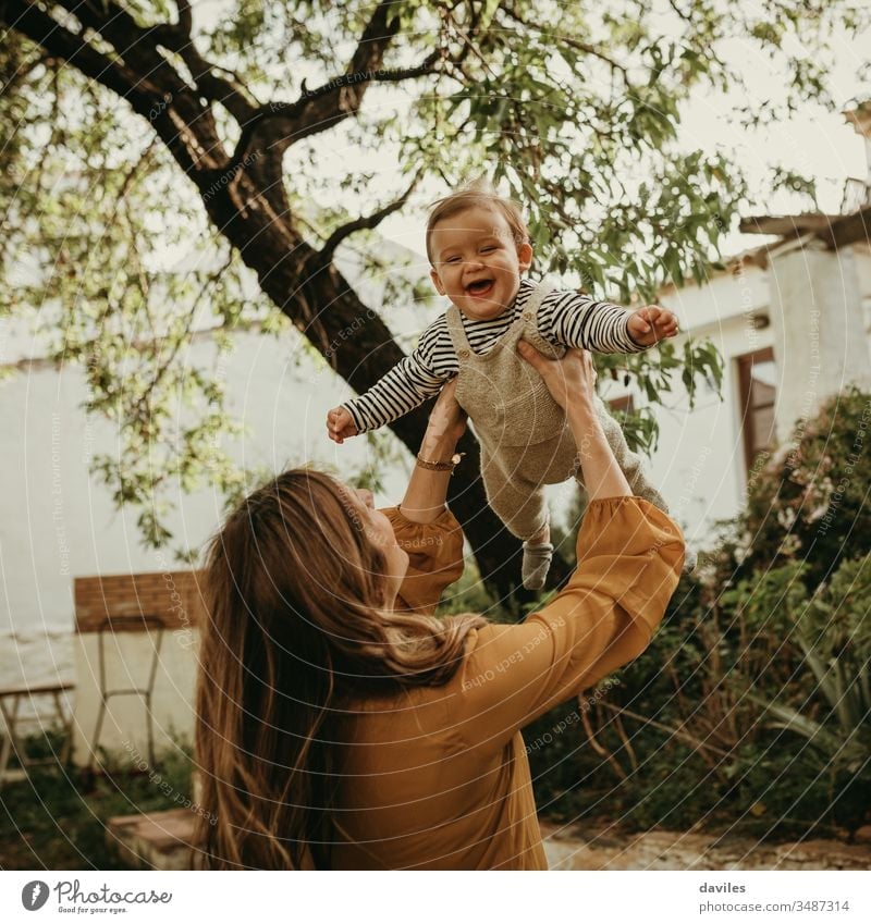 Mother holding her baby son up in the air, and baby laughing. playing adult fly arms throwing enjoying parenthood blond garden tree life woman newborn outdoors