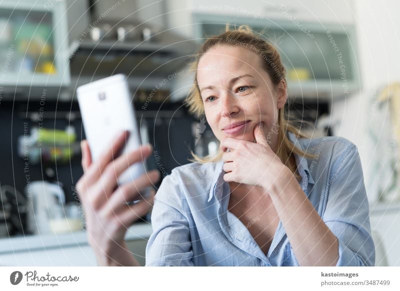 Young smiling cheerful pleased woman indoors at home kitchen using social media apps on mobile phone for chatting and stying connected with her loved ones. Stay at home, social distancing lifestyle.