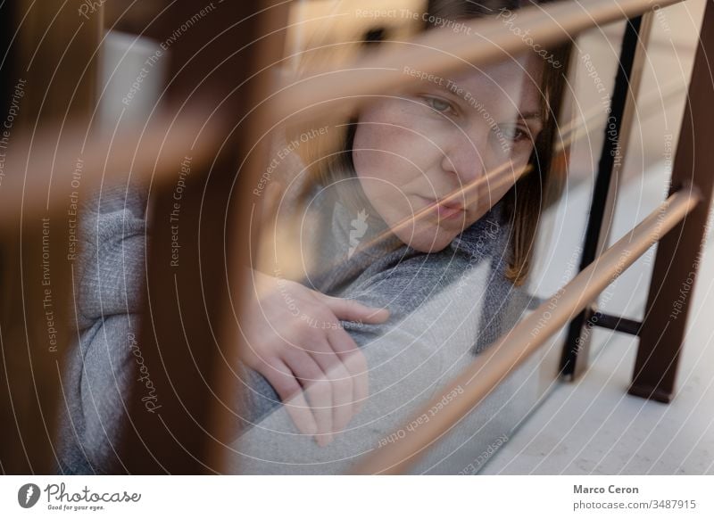 Beautiful Young woman looking melancholy through the glass to the street. Pensive sad girl, isolated at home during lock down by coronavirus. adult alone