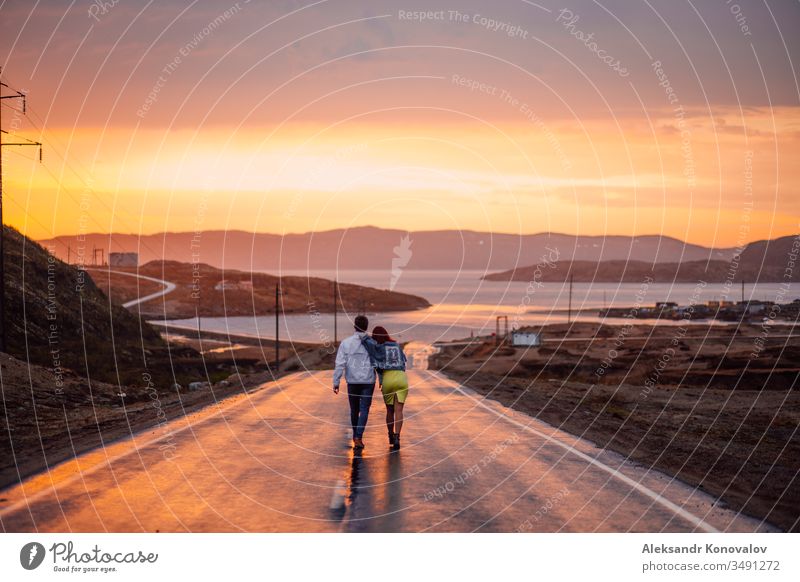 Young couple on the way to the Barents Sea on a wet road in a sunset Couple Sunset Golf Ocean Tundra polar Village Select Electricity pylon Asphalt Jeans
