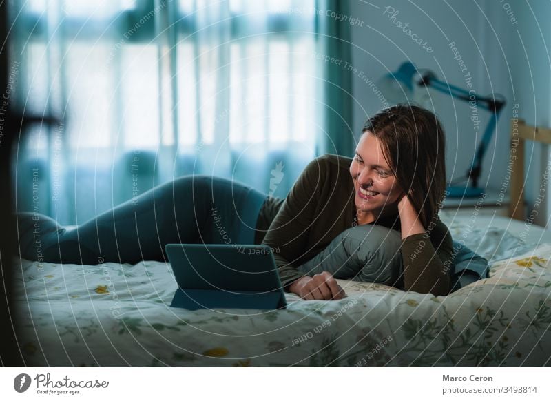 Smiling young woman lying on the bed while making a video call with her family. Attractive female using her digital tablet at home during quarantine. bedroom