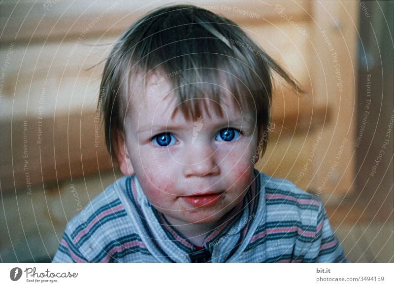 Small, sweet, curious toddler with a smudged mouth, blue eyes and red cheeks, looks curiously into the camera. Small, smudgy, cheeky, roll-eyed girl with striped shirt sits contentedly, happily at home. Happy childhood.
