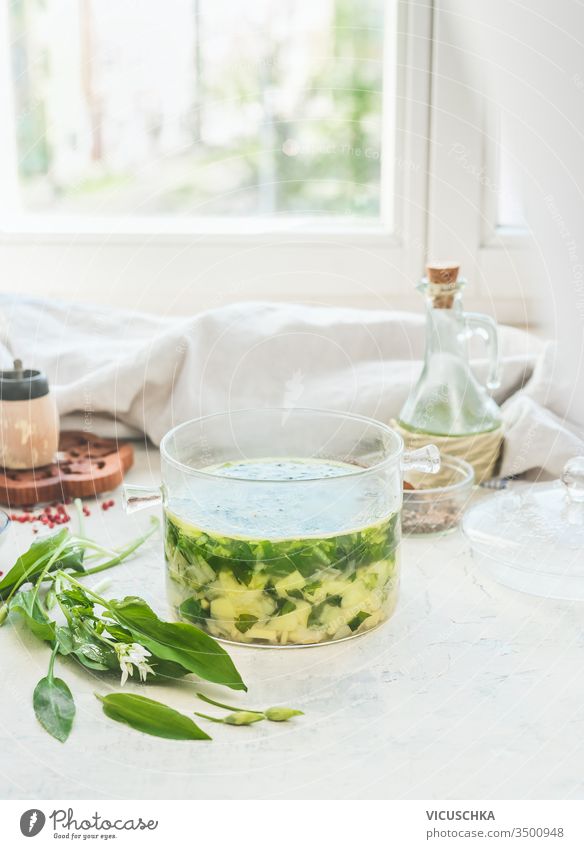 Green wild garlic soup in glass cooking pot stay on light table with ingredients at window background. Seasonal home cuisine. Vegan food. Healthy cooking and eating. Springtime recipes