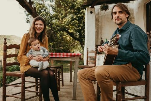 Beautiful couple with their baby infant enjoying sitting on the courtyard at countryside. Man is playing the guitar. Woman holds the baby. They look at the camera.
