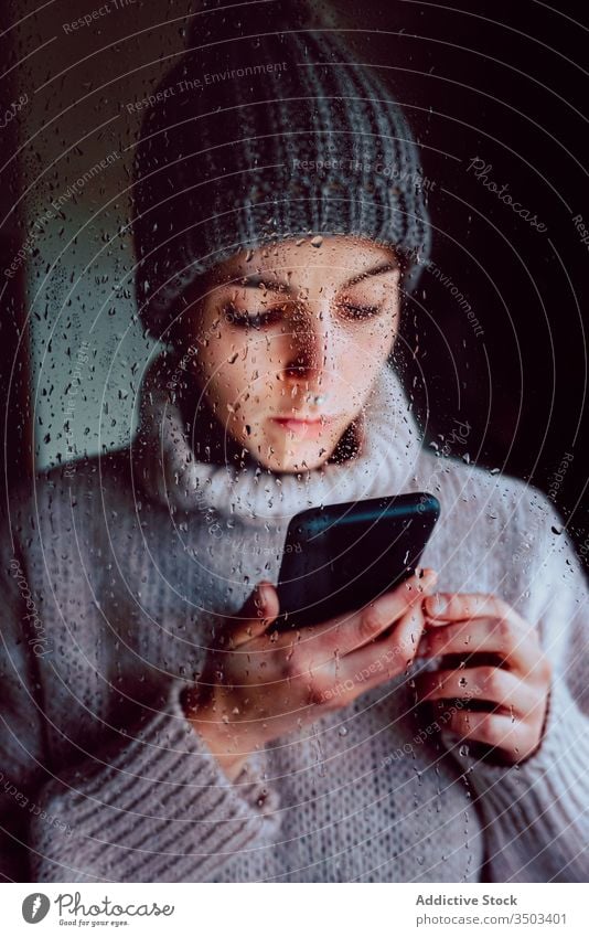 Young woman with smartphone standing near window home using lonely sad self isolation browsing gadget coronavirus covid quarantine young female casual sweater