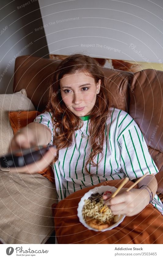 Young female eating noodles and watching TV woman ramen tv home lunch young rest japanese sofa film movie change channel remote control tradition authentic