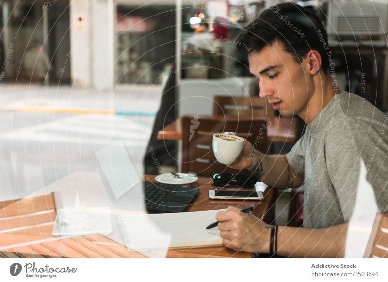Student drinking coffee and making notes in cafe man student write notebook young education male canteen cup table cafeteria window restaurant sit homework