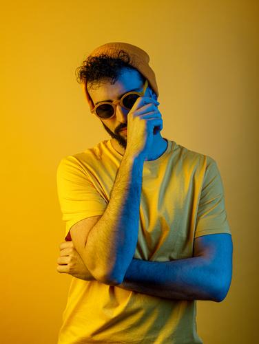Confident bearded male under yellow illumination man modern casual style touch face serious yellow light bright outfit trendy hat t shirt sunglasses apparel